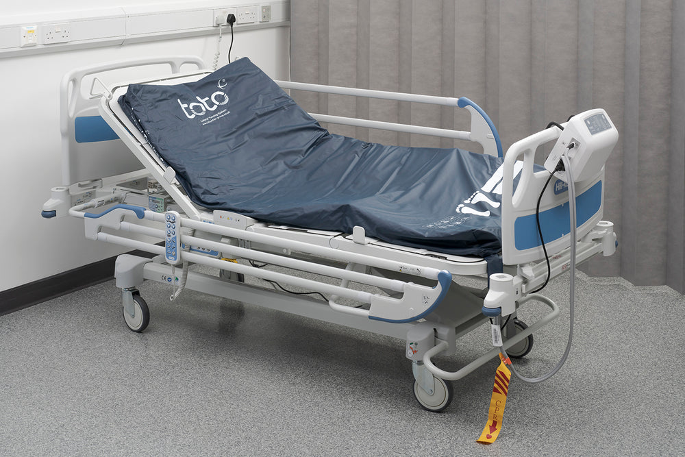 A hospital bed with the Toto platform in situ on the base of the bed, the control unit is hanging from the board at the foot of the bed, and the CPR tag is hanging from the tubing. The head of the bed is raised and Toto is in the tilted position.