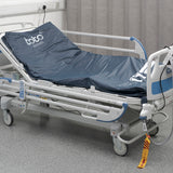 A hospital bed with the Toto platform in situ on the base of the bed, the control unit is hanging from the board at the foot of the bed, and the CPR tag is hanging from the tubing. The head of the bed is raised and Toto is in the tilted position.